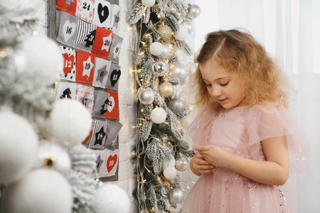 Toddler child gets new advent calendar task, waiting for Christmas. Festive mood for cute curly girl. Kid gets gift from advent pocket with dates from the wall at home.の写真素材