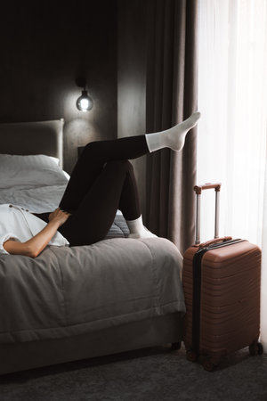 Woman sits with her legs on a suitcase, showing sense of solitude, having rest. Female in a hotel room, reflecting on her travel experience, feeling a wave of reliefの写真素材