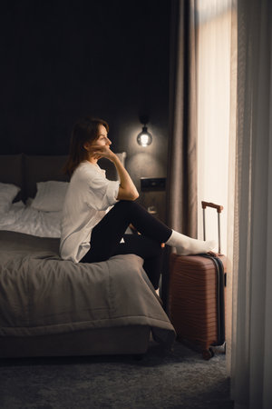 Woman sits on the edge of cozy comfortable hotel bed, looking out the window dreaming. Person reflecting on travel experiences and plans, surrounded by her luggage.の写真素材