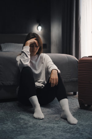 Depressed woman sitting in hotel room with her suitcase nearby. Female emotional crisis during solo travel, feeling sense of loneliness, escape from complicated situationの写真素材