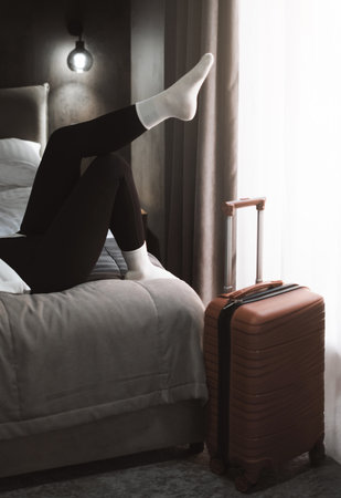 Woman sits with her legs on a suitcase, showing sense of solitude, having rest. Female in a hotel room, reflecting on her travel experience, feeling a wave of reliefの写真素材