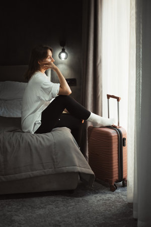 Woman sits on the edge of cozy comfortable hotel bed, looking out the window dreaming. Person reflecting on travel experiences and plans, surrounded by her luggage.の写真素材