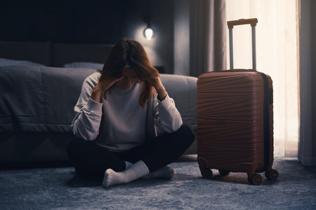Sad woman sits on the floor, feeling overwhelmed and lonely. Female with suitcase having frustration. Dark hotel room with curtains, griefの写真素材