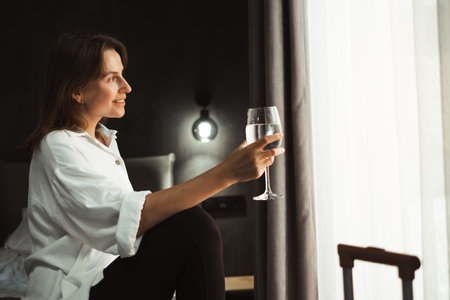 Woman sits on the edge of cozy comfortable hotel bed, looking out the window dreaming with glass of water. Person reflecting on travel experiences and plans, surrounded by her luggage.の写真素材
