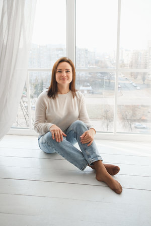 woman sits comfortably on the floor by a large window, enjoying the calmness of her home. Soft light fills the room, tranquil atmosphere perfect for relaxation. Portraitの写真素材