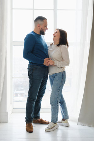 Loving couple at 40s stands by large window in home, enjoying quality time together. Family smiles, hug, moment of joy and togethernessの写真素材