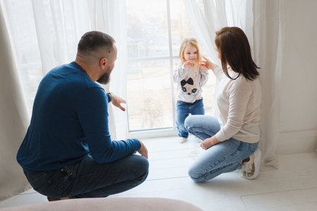 Parents crouch down to engage with a little girl who stands by a window in cozy, bright living room filled with warmth and love. Connection and fun time with familyの写真素材