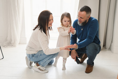 Family engaged in a fun activity in bright living room. Toddler child sits on her fathers shoulders while the mother smiles and reaches out, creating a loving atmosphere.の写真素材