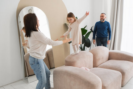Mother encourages her daughter to balance on soft furniture in a bright living room. Father observes with pride, showcasing the warmth and love of family interactions and fun timeの写真素材
