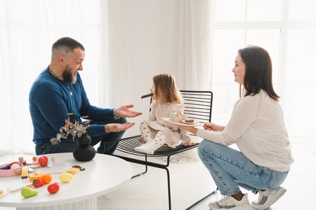 Parents crouch down to engage with a little girl who stands by a window in cozy, bright living room filled with warmth and love. Connection and fun time with familyの写真素材