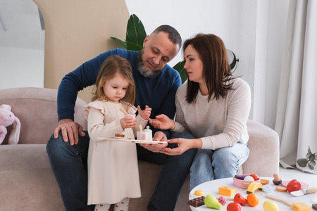 Parents crouch down to engage with a little girl who stands by a window in cozy, bright living room filled with warmth and love. Connection and fun time with familyの写真素材