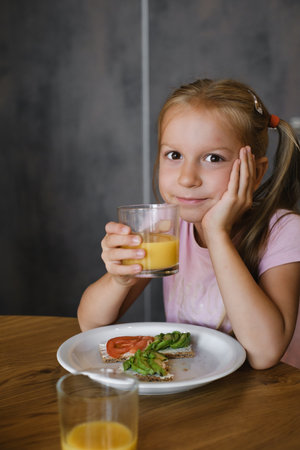 young girl smiles drinking glass of juice and enjoys whole grain toast topped with fresh tomatoes and avocado.の写真素材