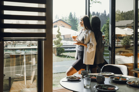 Relaxing in a modern hotel dining area with scenic view. Woman traveler enjoys drink while looking out of large window in a contemporary hotel. Table is set with delicious meal, cozy atmosphereの写真素材