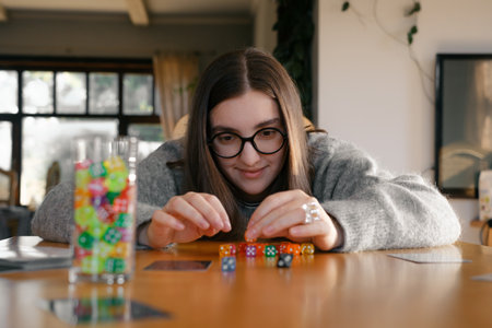 Young woman focuses intently while stacking colorful dice on wooden table. Excitement and joy during table game night. Colorful dice make female concentratedの写真素材