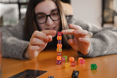 Young woman focuses intently while stacking colorful dice on wooden table. Excitement and joy during table game night. Colorful dice make female concentratedの写真素材