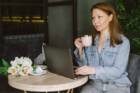 Woman working on laptop in cafe. Middle-aged freelancer with cup of coffee typing and reading. Loft working place with green plantsの写真素材