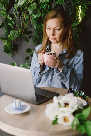 Woman working on laptop in cafe. Middle-aged freelancer with cup of coffee typing and reading. Loft working place with green plantsの写真素材