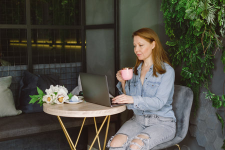 Woman working on laptop in cafe. Middle-aged freelancer with cup of coffee typing and reading. Loft working place with green plantsの写真素材