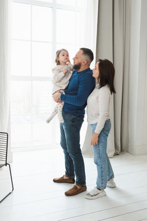 Family engaged in a fun activity in bright living room. Toddler child sits on her fathers shoulders while the mother smiles and reaches out, creating a loving atmosphere.の写真素材