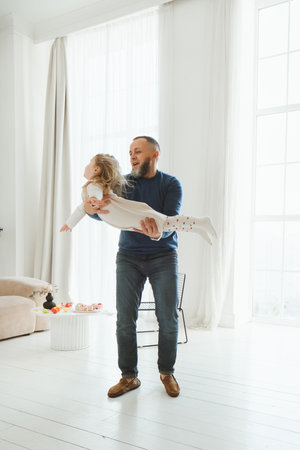 Father lifts his daughter joyfully in bright and airy living room. Large windows, creating a warm and loving atmosphere. Active game and parenting. Fathers dayの写真素材