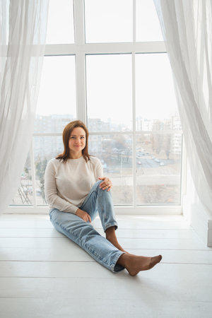woman sits comfortably on the floor by a large window, enjoying the calmness of her home. Soft light fills the room, tranquil atmosphere perfect for relaxation. Portraitの写真素材