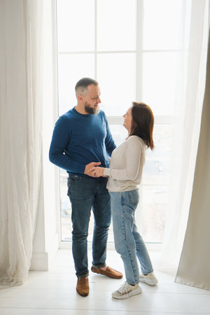 Loving couple at 40s stands by large window in home, enjoying quality time together. Family smiles, hug, moment of joy and togethernessの写真素材