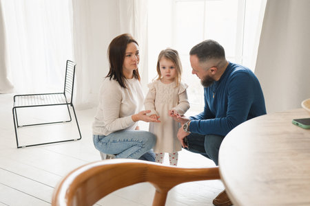 Parents crouch down to engage with a little girl who stands by a window in cozy, bright living room filled with warmth and love. Connection and fun time with familyの写真素材