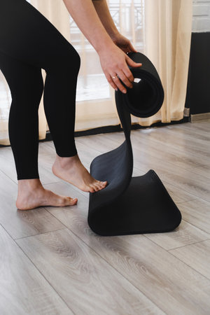 Rolling Black yoga mat in fitness studio. Closeup of a person getting ready for balance, flexibility sport activity for adults in gym. Woman hands collecting equipmentの写真素材