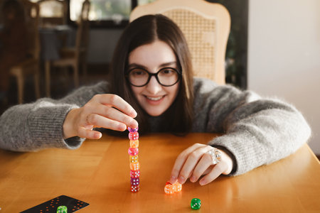 Young woman focuses intently while stacking colorful dice on wooden table. Excitement and joy during table game night. Colorful dice make female concentratedの写真素材
