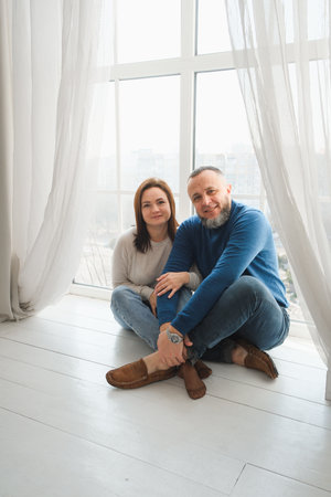 Couple sit closely together by sunny windows, enjoying tender moments filled with love and connection. Warmth of family time at home.の写真素材