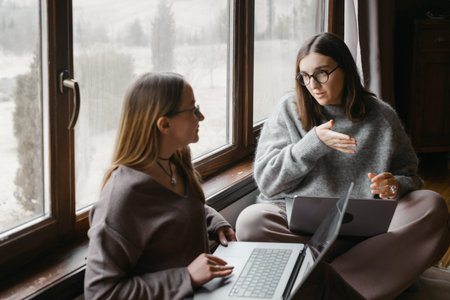 Female freelancers are sitting near large window, working on their laptop.