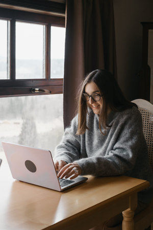 Woman content creator using a laptop. Female in cozy sweater smiling and consulting on-line. Remote...