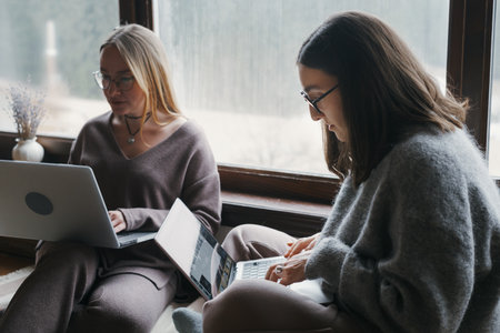 Two women are working on their laptops near a bright window. They are wearing comfortable clothes and working from their apartment on computers. The women appear focused on their screens.の写真素材