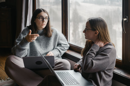 Two Young Women Discussing Work Project Sitting Near Window with Laptops