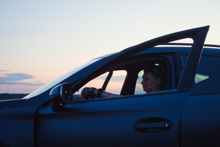 Man sitting in car driver seat with open door at sunset