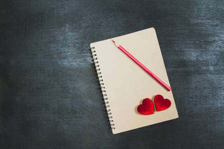Two red hearts and red color pencil on brown notebook on black wooden background, dark tone, valentine concept.の写真素材