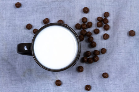 Top view of milk in black ceramic cup and chocolate cereals on white cloth background with copy...