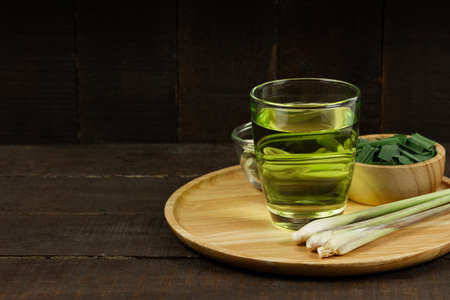 Lemon grass tea in glass and fresh lemon grass and green leaf in wooden bowl on wooden background. Healthy herbal drink concept dark tone.の写真素材