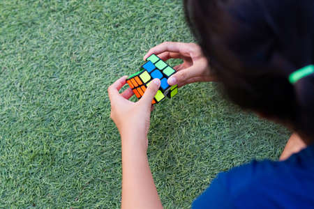 SAMUT-SAKHON, THAILAND - APRIL 02, 2021 : Hands of a young girl trying to solve the Rubik's Cube puzzle. She sitting on the grass field.のeditorial素材