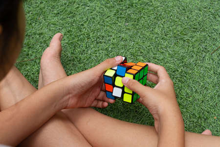 SAMUT-SAKHON, THAILAND - APRIL 04, 2021 : Hands of a young girl trying to solve the Rubik's Cube puzzle. She sitting on the grass field.のeditorial素材