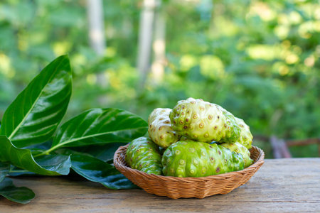Noni fruit or Morinda Citrifolia on Wicker basket and green leaf on wooden table with blur...