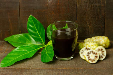 Noni juice in transparent glass and noni fruit with green leaf on wooden background.の写真素材