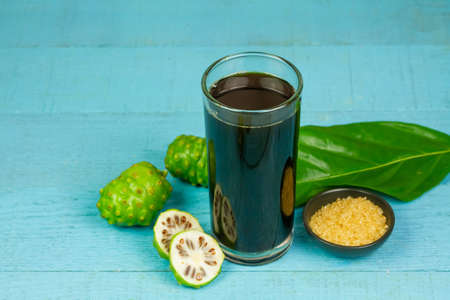 Noni juice in transparent glass and fresh noni fruit with green leaf on blue wooden background.の写真素材