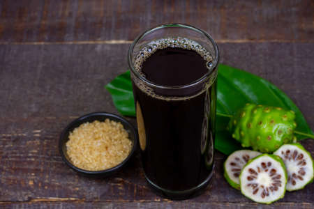 Noni juice in transparent glass cup and fresh noni fruit with green leaf on rustic wooden background.の写真素材