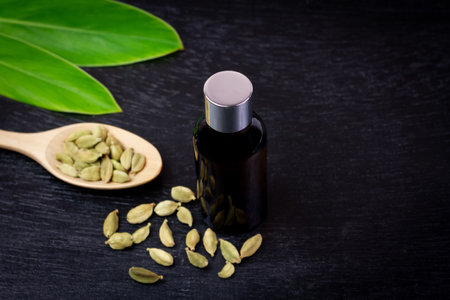 Cardamom essential oil in glass bottle with dry cardamom spice on wooden spoon and green leaf on black wooden background.の写真素材
