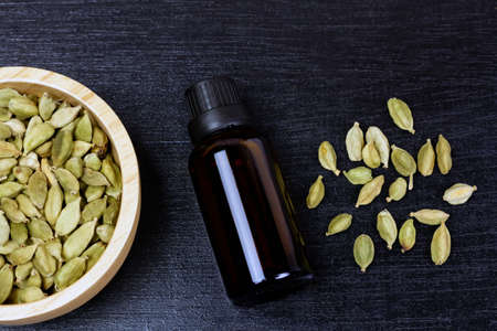 Cardamom essential oil in glass bottle with dry cardamom spice on wooden bowl on black wooden background, top view.の写真素材