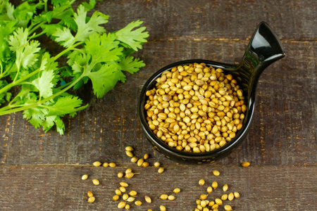Coriander seeds on black bowl with green leaf on rustic wooden background, top view. Coriander seeds...