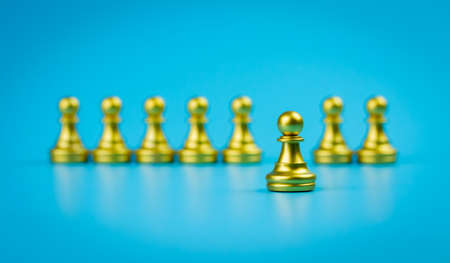 Single gold pawn chess figure stands in front of multiple gold pawn chess figure on blue background, leadership concept.の写真素材