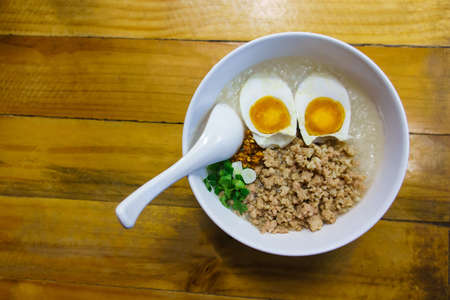 Minced pork congee with egg, fresh ginger and  coriander on white cup and spoon over the wood table.の写真素材