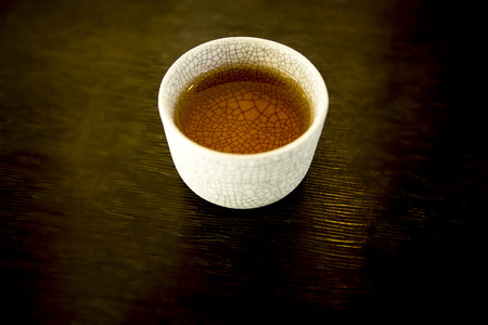 Fleshing delicious welcome drink water in small pattern decoration ceramic glass on the dark brown pattern background.Drink water color is clear brown.Can be put tea inside.Actually it is apple juice.の写真素材